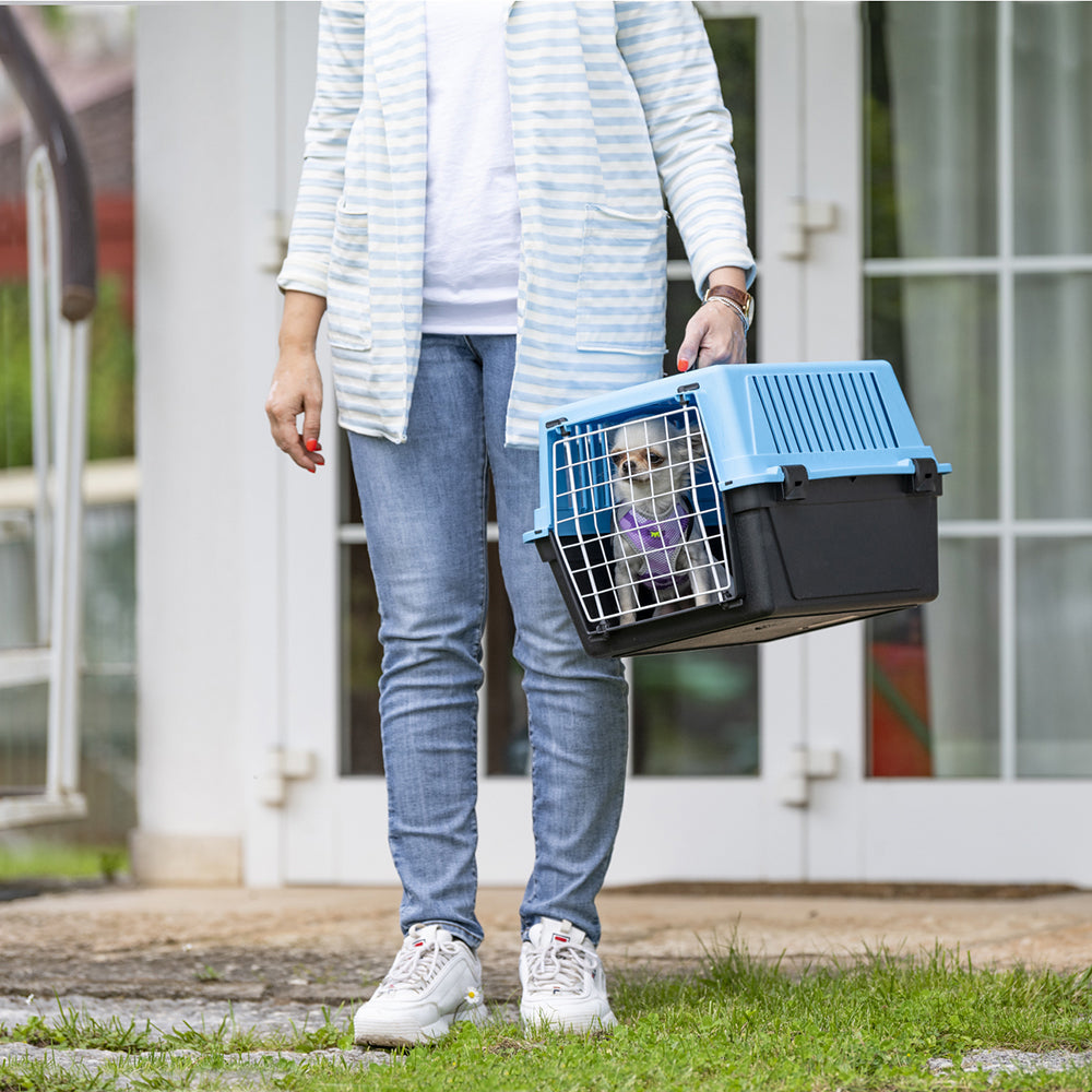Personne tenant une cage de transport pour animaux avec un chien à l'intérieur, devant une maison. Transporteur bleu et noir, idéal pour voyages sécurisés.