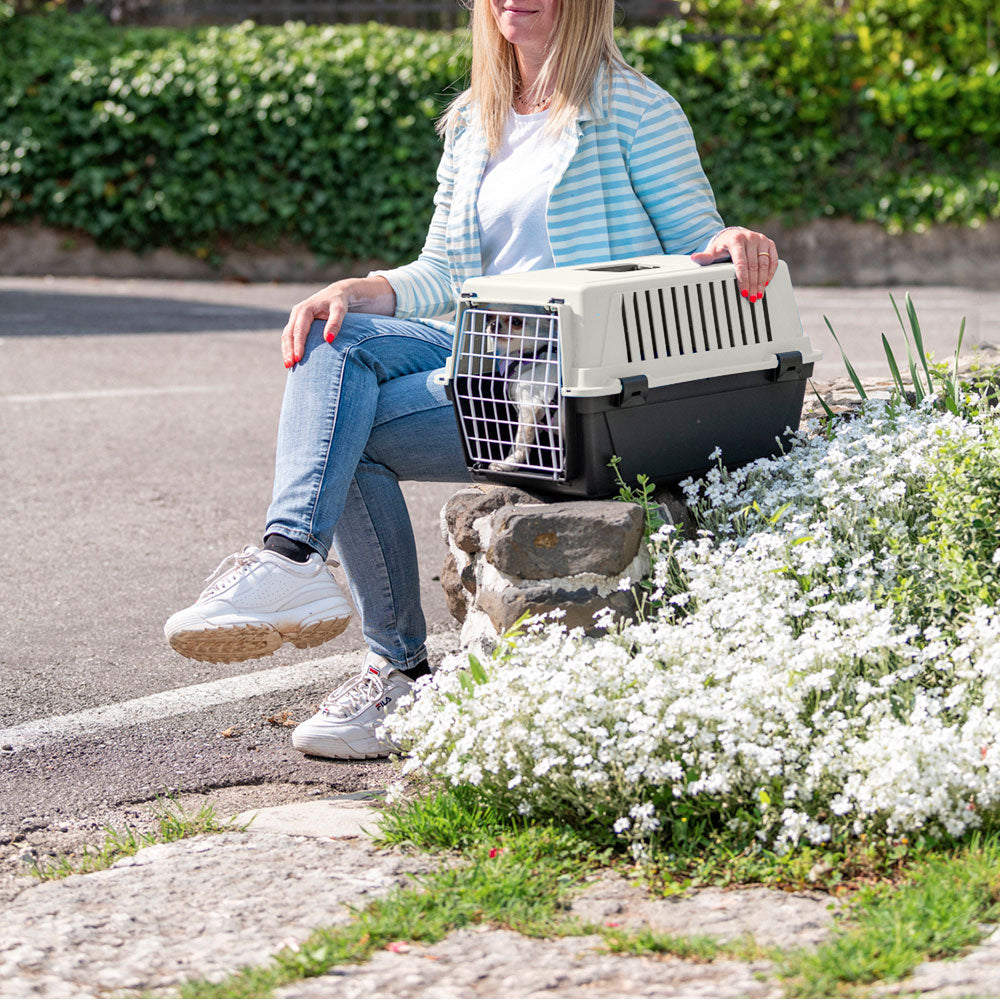Femme assise avec une cage de transport pour chat, entourée de fleurs blanches. Accessoire de voyage pour animaux, extérieur, printemps.